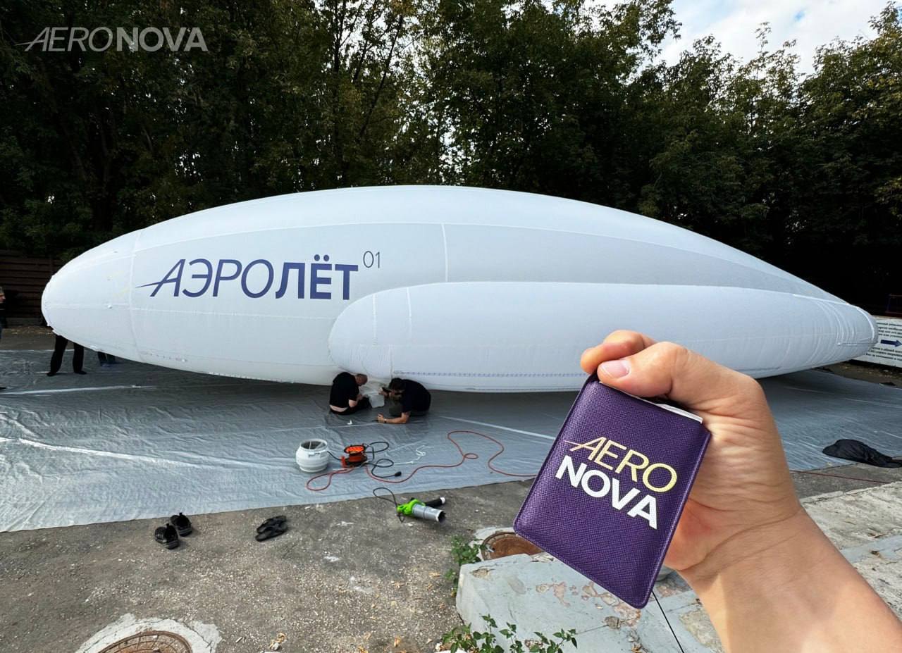 A real AERONOVA airship inflated outdoors, with an AERONOVA brand card held up in the foreground.